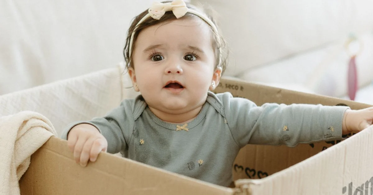 Joli bébé avec un bandeau assise dans une boîte en carton, affichant une expression curieuse.
