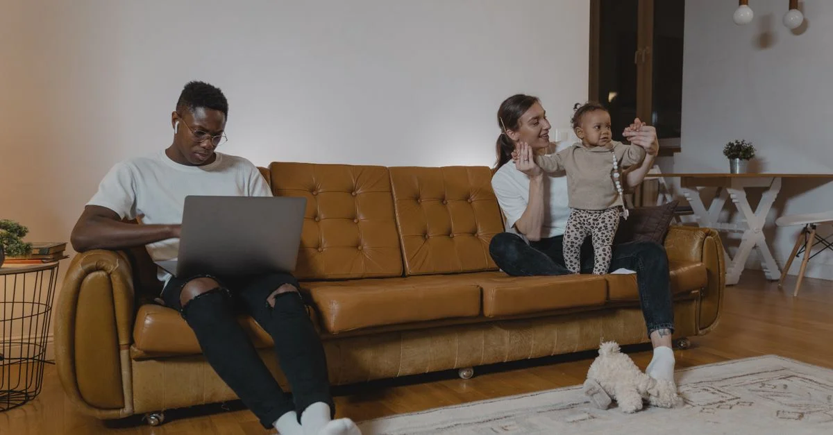 Une famille passe du temps ensemble sur le canapé, avec un homme travaillant sur un ordinateur portable et une femme jouant avec un bébé.