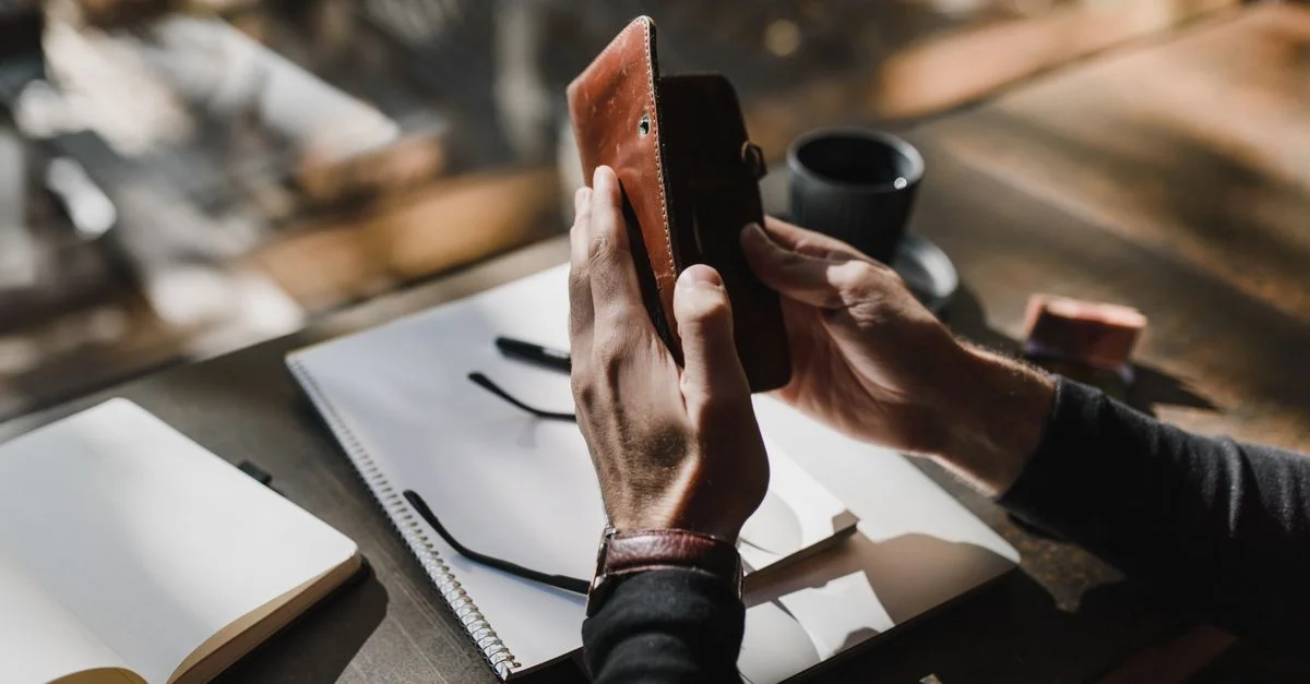 Un gros plan des mains d'un homme tenant un portefeuille en cuir au-dessus d'un cahier sur une table en bois.